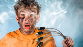 A boy covered in grease with a shocked look on his face holding 4 cables while smoke rises behind him on a blue background